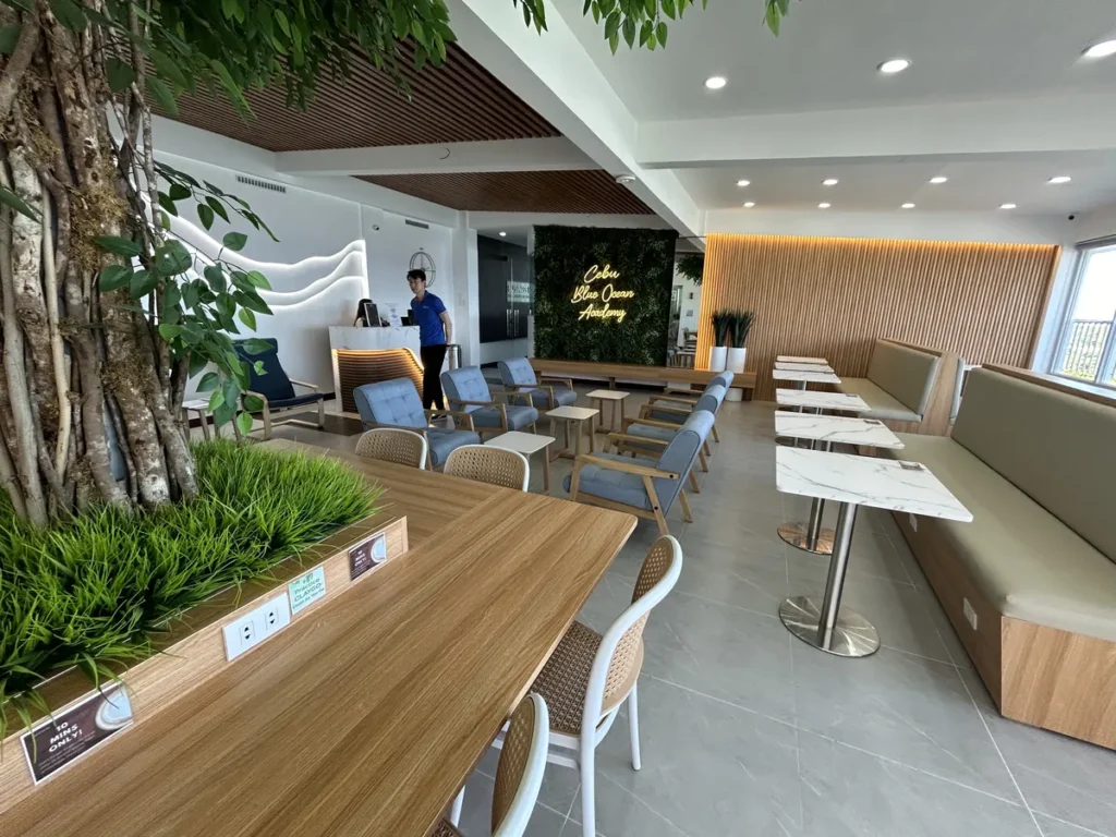 필리핀 세부 블루오션(BLUEOCEAN) 어학원 라운지로 인조나무와 대리석 테이블, 안쪽 데스크가 보이는 밝은 학습 휴식 공간입니다