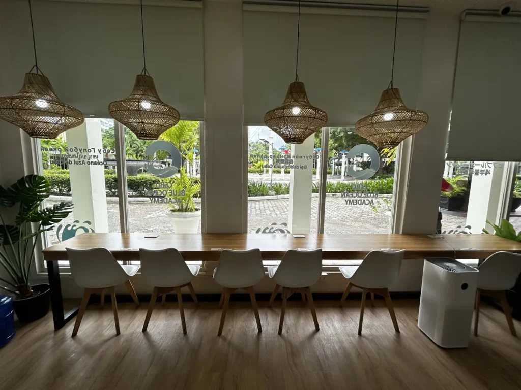필리핀 세부 블루오션(BLUEOCEAN) 어학원 라운지로 라탄 펜던트 조명 아래 긴 우드 테이블과 흰 의자가 놓인 창가 좌석 공간입니다