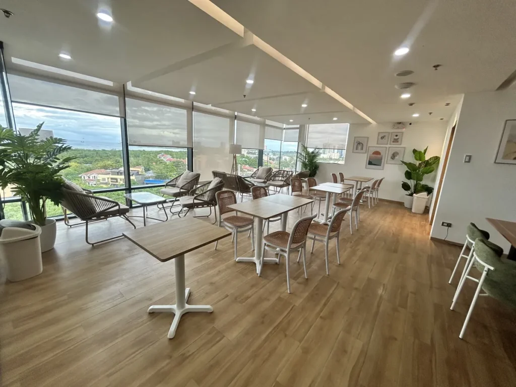 필리핀 세부 블루오션(BLUEOCEAN) 어학원 외부숙소 부대시설로, 통유리창 너머 바다와 시내 전망이 보이는 라탄 의자와 원목 테이블의 스카이라운지 휴게공간입니다.
