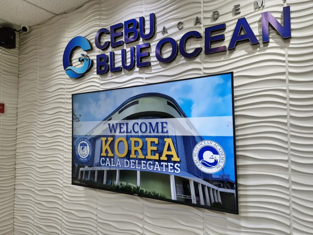 필리핀 세부 블루오션(BLUEOCEAN) 어학원 입구홀 웨이브 벽면에 부착된 금색 CEBU BLUE OCEAN 로고와 환영 영상 모니터