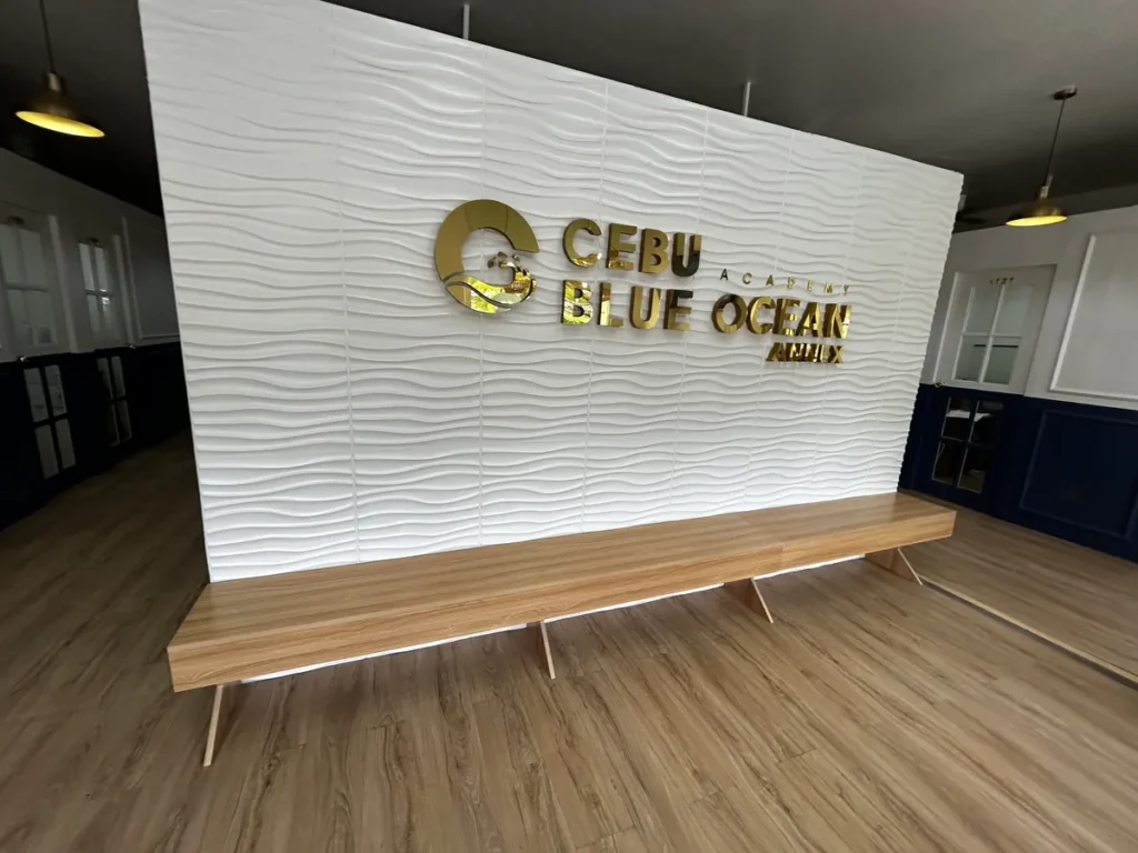 필리핀 세부 블루오션(BLUEOCEAN) 어학원 입구홀 흰 웨이브 벽면의 금색 BLUE OCEAN 로고와 원목 긴 벤치, 우드 바닥 라운지