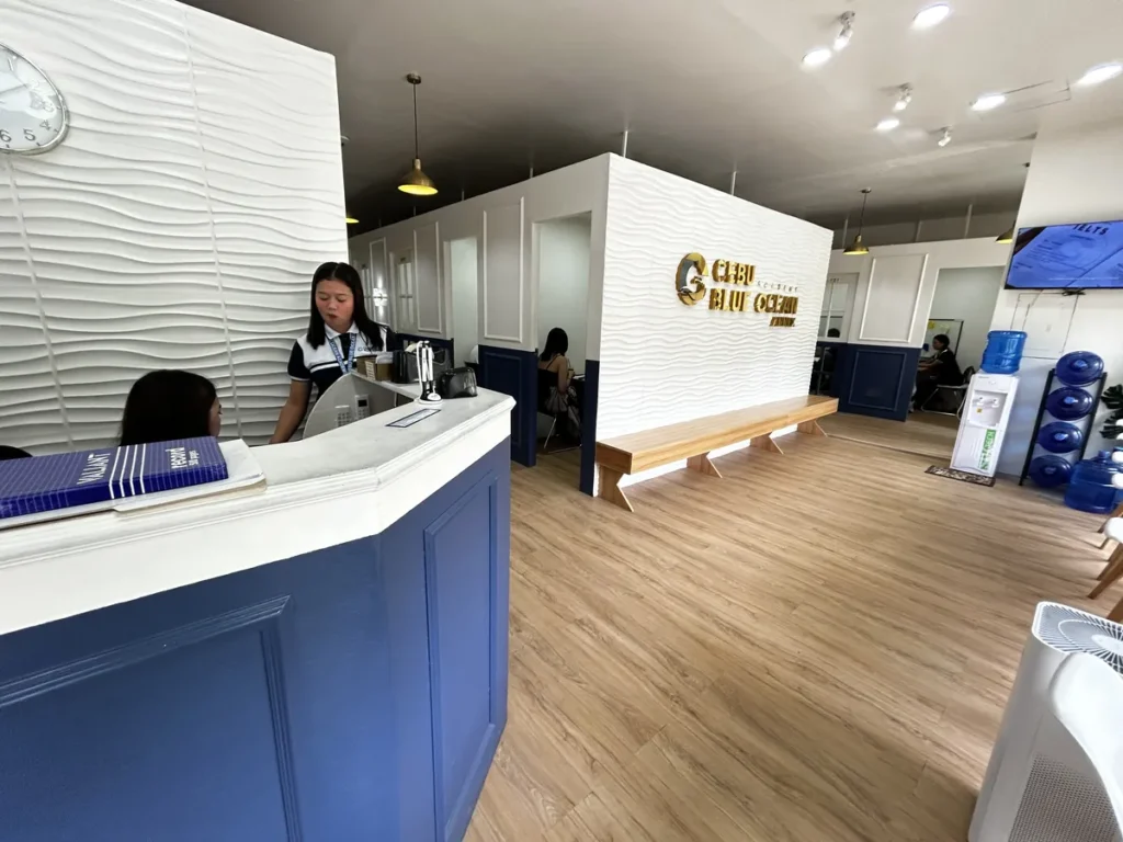 필리핀 세부 블루오션(BLUEOCEAN) 어학원 입구홀에서 직원이 학생을 응대하는 안내 데스크와 금색 로고 벽, 정수기 코너
