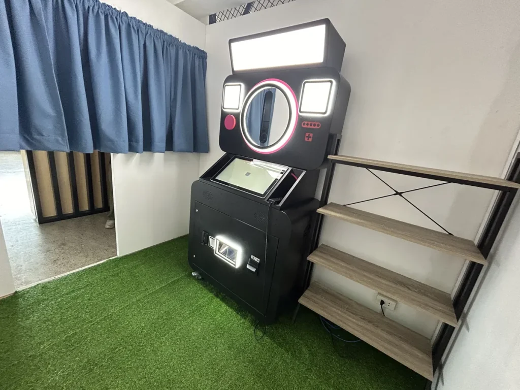 필리핀 세부 CIA어학원 인생네컷 포토부스, 파란 커튼 옆 인조잔디 바닥에 놓인 카메라 모양 즉석 사진 기기와 선반