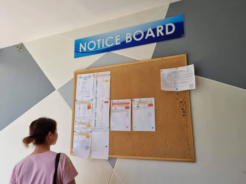 필리핀 세부 CIA어학원 부대시설 안내판으로, NOTICE BOARD 사인 아래 시험 일정과 여행 신청서 등 안내문이 붙은 코르크 게시판을 학생이 보고 있습니다.