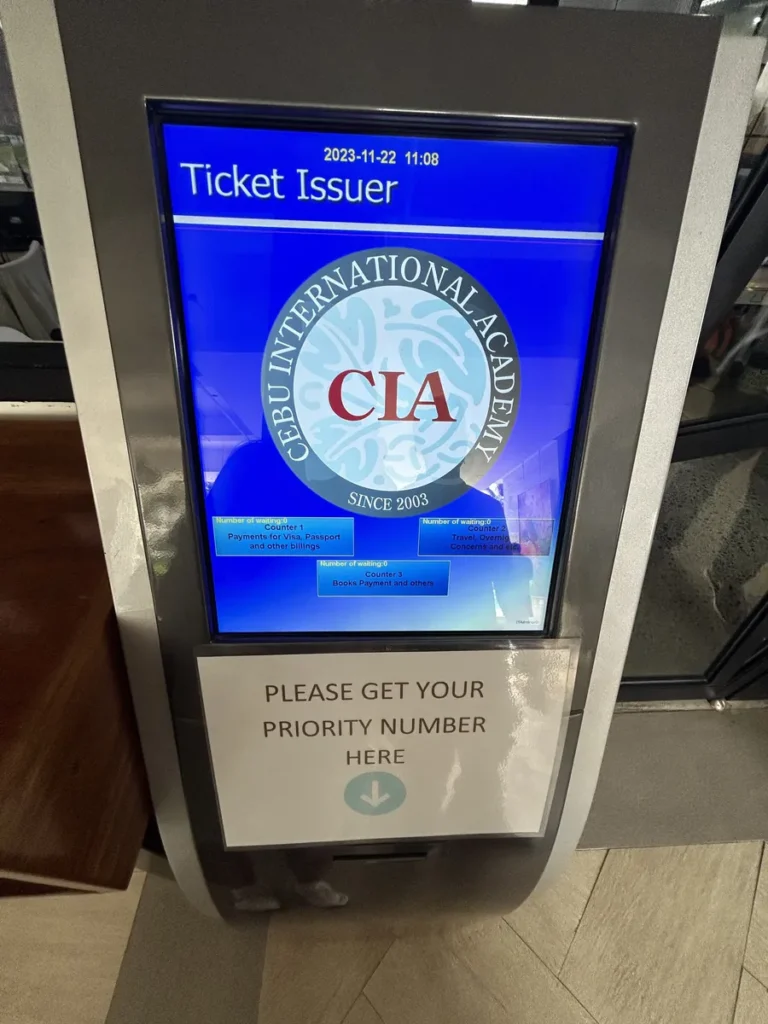 필리핀 세부 CIA어학원 부대시설 안내판으로, 카운터별 안내가 표시된 Ticket Issuer 번호표 발권기와 PLEASE GET YOUR PRIORITY NUMBER 안내문이 보입니다.