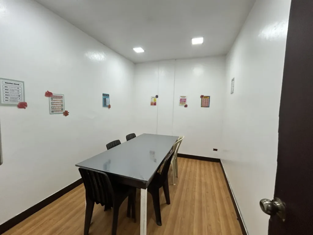 필리핀 클락 벨라(BELA) 어학원 그룹강의실로, 회색 큰 테이블에 의자 여러 개가 둘러싸고 원목 무늬 바닥과 영어 학습 포스터가 붙은 빈 교실입니다.