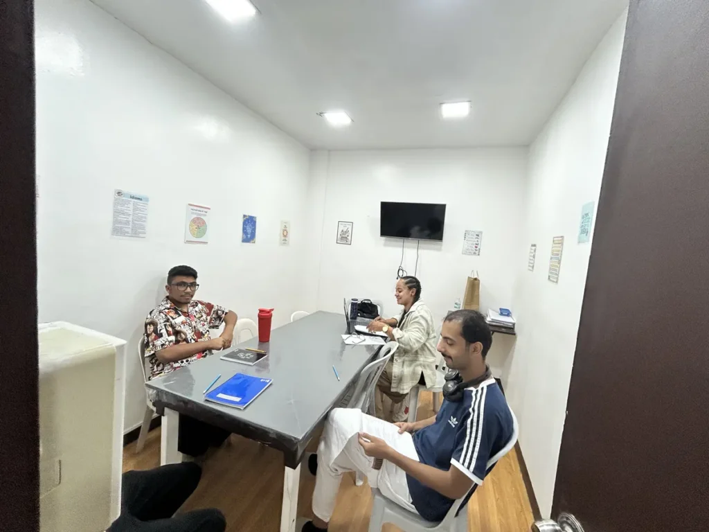 필리핀 클락 벨라(BELA) 어학원 그룹강의실로, 벽걸이 TV와 영어 포스터가 붙은 벽 아래 학생 세 명이 테이블에 둘러앉아 공부하는 모습입니다.