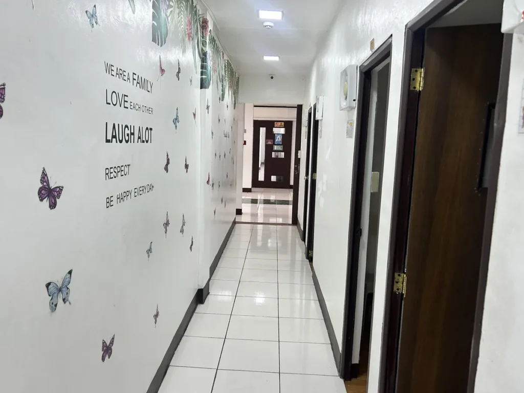 필리핀 클락 벨라(BELA) 어학원 강의실 복도로, 나비 스티커와 열대 잎 그래픽 벽화 아래 격려 문구가 적힌 복도를 따라 강의실 문들이 늘어선 모습입니다.