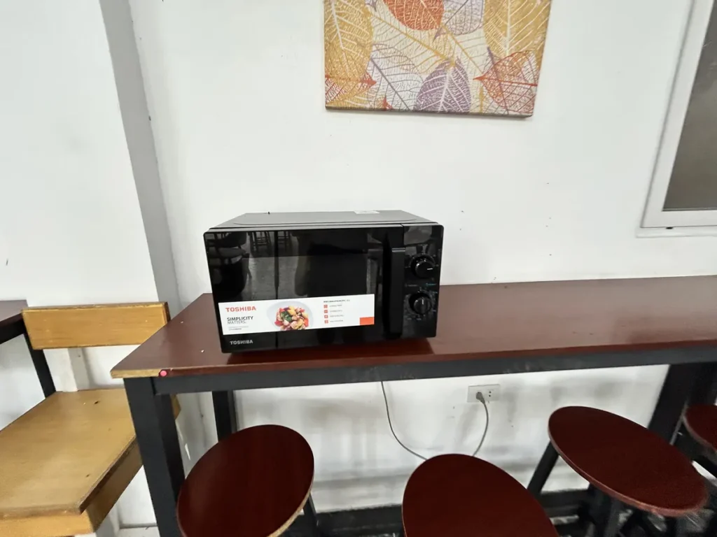 필리핀 클락 벨라(BELA) 어학원 부대시설 매점으로, 원목 바 테이블 위에 놓인 토시바 전자레인지와 그 아래 스툴이 보이는 모습입니다.