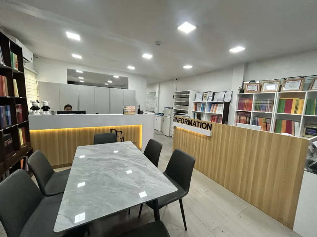 필리핀 클락 벨라(BELA) 어학원 부대시설 오피스로, 원목 책장과 대리석 테이블·의자, 나무 패널의 리셉션 데스크가 있는 사무 공간입니다.