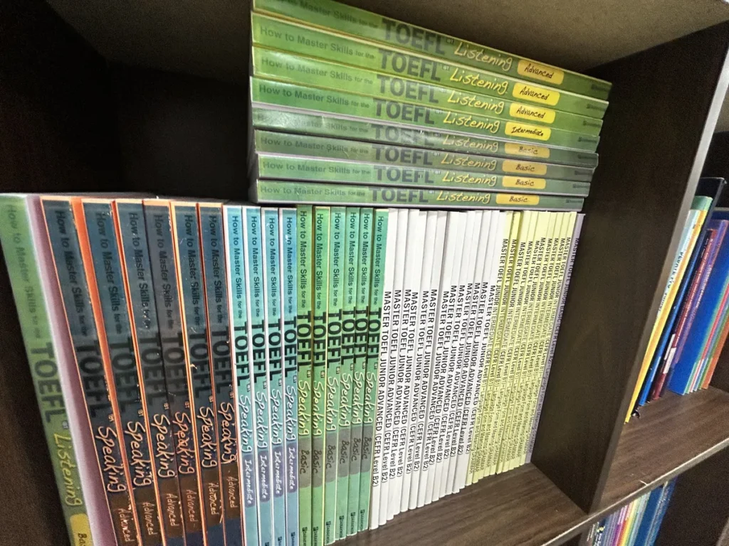 필리핀 클락 벨라(BELA) 어학원 부대시설 오피스로, TOEFL 리스닝·스피킹과 TOEFL 주니어 교재가 나란히 꽂힌 책장을 가까이서 찍은 모습입니다.