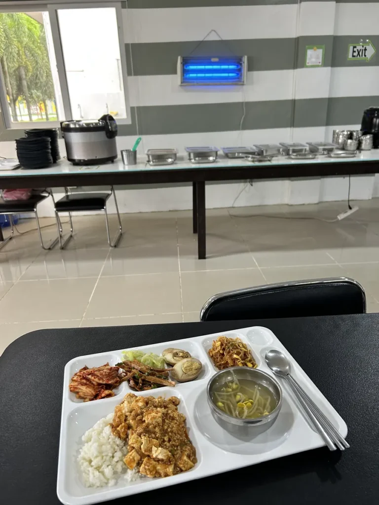 필리핀 클락 벨라(BELA) 어학원 식당 식사로, 모기퇴치램프와 Exit 표지판이 보이는 식당 테이블 위에 밥과 반찬이 담긴 식판이 놓여 있습니다.