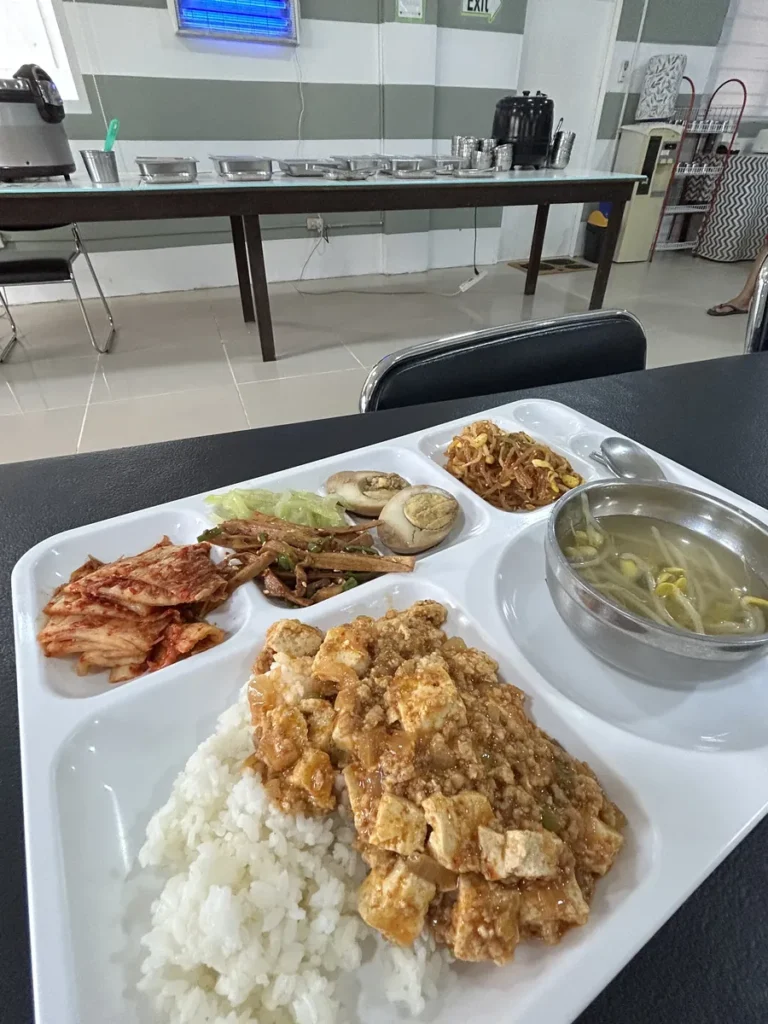 필리핀 클락 벨라(BELA) 어학원 식당 식사로, 줄무늬 벽과 창문이 있는 식당 안쪽 배식대 앞 테이블에 반찬과 국이 담긴 식사 식판이 놓여 있습니다.