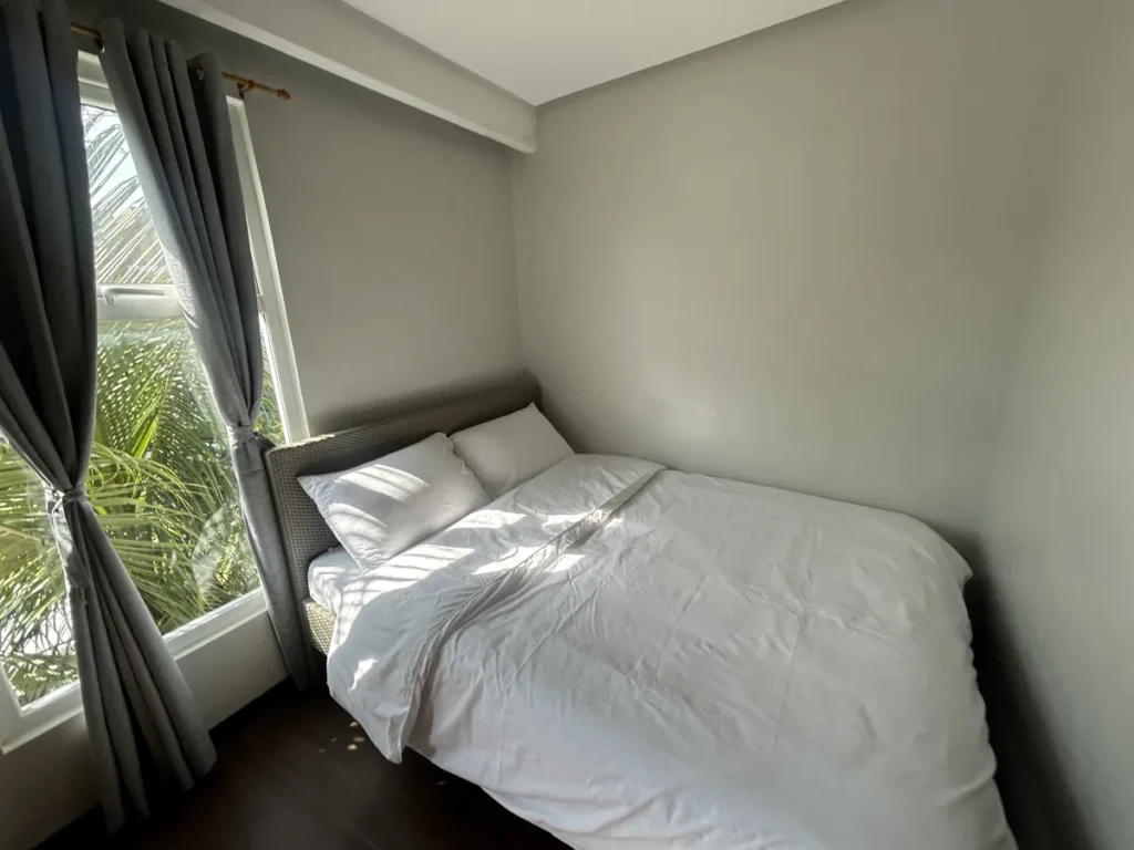 필리핀 세부 바나나키즈(BK) 어학원 외부 독채 기숙사 침실로, 회색 커튼 창가에 야자수 잎이 비치고 흰색 침구 더블베드가 붙어 있습니다.