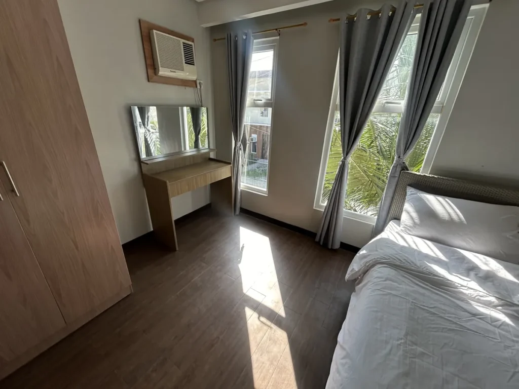 필리핀 세부 바나나키즈(BK) 어학원 외부 독채 기숙사 침실로, 거울 달린 화장대와 벽걸이 에어컨 옆 창밖으로 이웃 건물과 야자수가 내다보입니다.