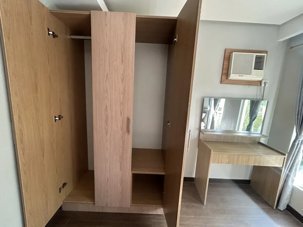 필리핀 세부 바나나키즈(BK) 어학원 외부 독채 기숙사 침실로, 문을 연 우드톤 옷장 내부 옆으로 거울 화장대와 벽걸이 에어컨이 놓인 수납 구성입니다.