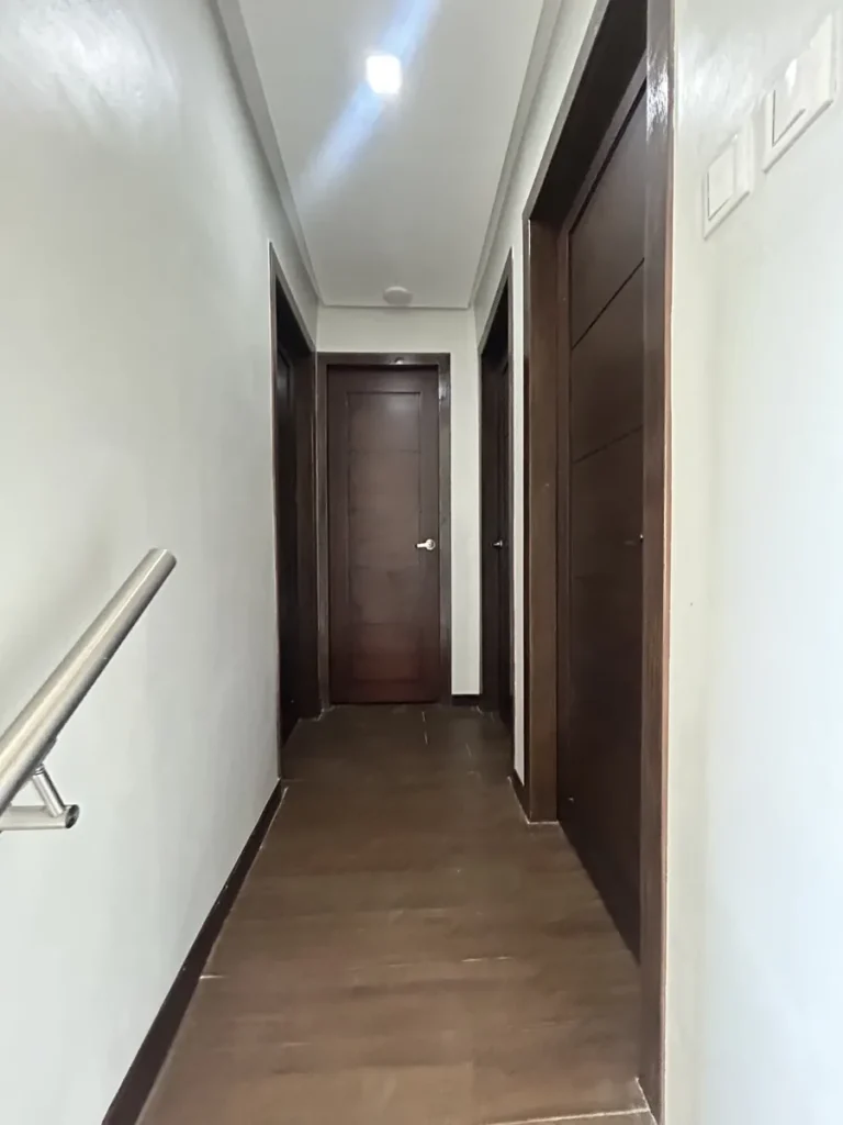 필리핀 세부 바나나키즈(BK) 어학원 외부 독채 기숙사 복도로, 짙은 나무문 두 개가 마주한 좁은 통로에 우드톤 바닥과 금속 난간 일부가 보입니다.