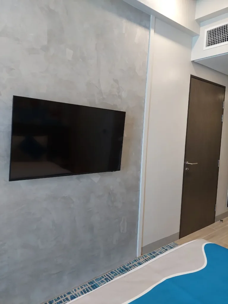 필리핀 세부 바나나키즈(BK) 어학원 외부 호텔 기숙사 객실 TV로, 회색 마감 벽면에 걸린 벽걸이 TV와 짙은 색 원목 출입문이 나란한 모습입니다.
