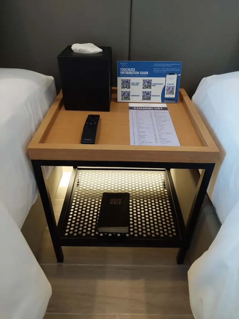 필리핀 세부 바나나키즈(BK) 어학원 외부 호텔 기숙사 침대 협탁으로, 리모컨과 채널 안내지 아래 칸에 성경책이 놓인 두 침대 사이 수납 테이블입니다.