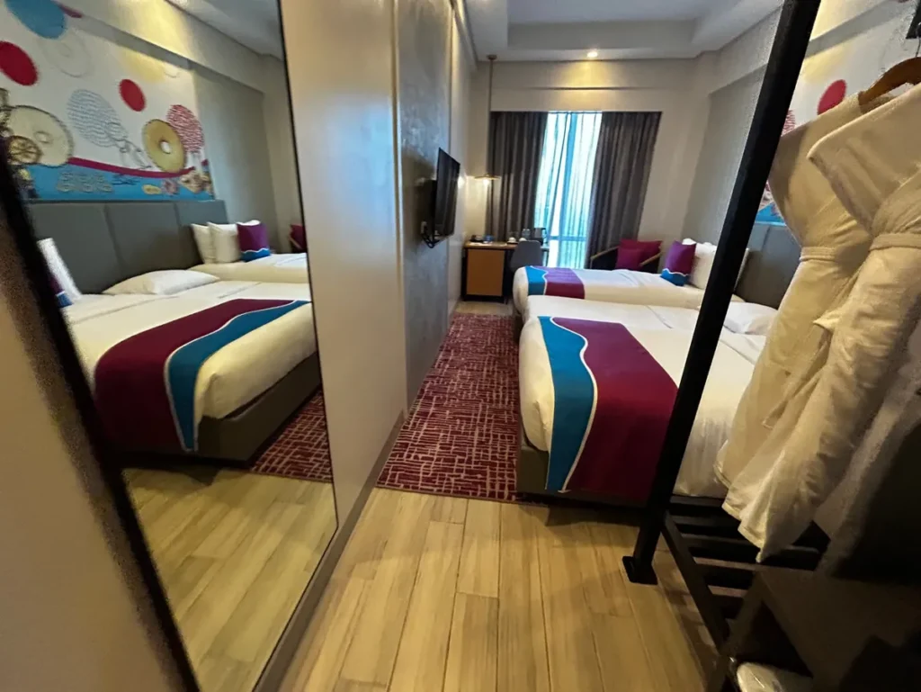 필리핀 세부 바나나키즈(BK) 어학원 외부 호텔 기숙사 트윈 객실로, 큰 거울에 침대와 벽화가 비치고 옷걸이에는 흰 가운이 걸려 있습니다.