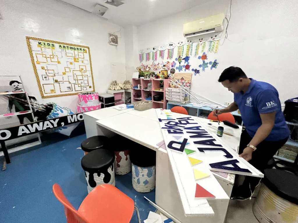 필리핀 세부 바나나키즈(BK) 어학원 키즈룸 미술공작실로, KIDS ARTWORKS 게시판 앞에서 직원이 알파벳 조형물을 칠하는 모습입니다.