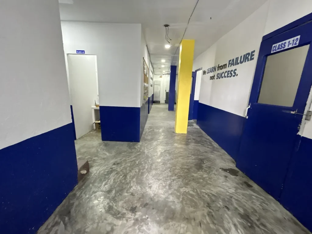 필리핀 세부 바나나키즈(BK) 어학원 강의실 복도로, 노란 기둥과 게시판을 지나 CLASS 1-12 문까지 이어지는 콘크리트 바닥 통로입니다.