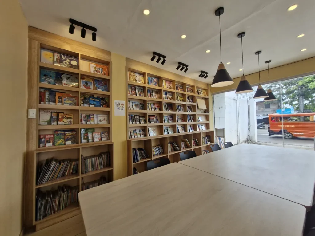 필리핀 세부 바나나키즈(BK) 어학원 부대시설 영어도서관으로, 펜던트 조명 아래 긴 학습 테이블과 양옆 책장이 늘어선 열람실 모습입니다.