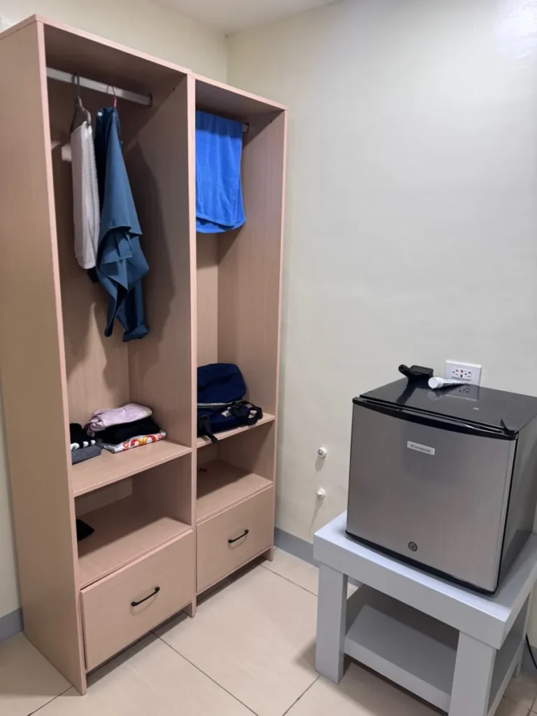 필리핀 세부 셀라(CELLA) 유니캠퍼스 어학원 기숙사 1인실로, 옷걸이와 서랍이 있는 원목 옷장과 그 옆 미니 냉장고가 놓인 수납 공간입니다.