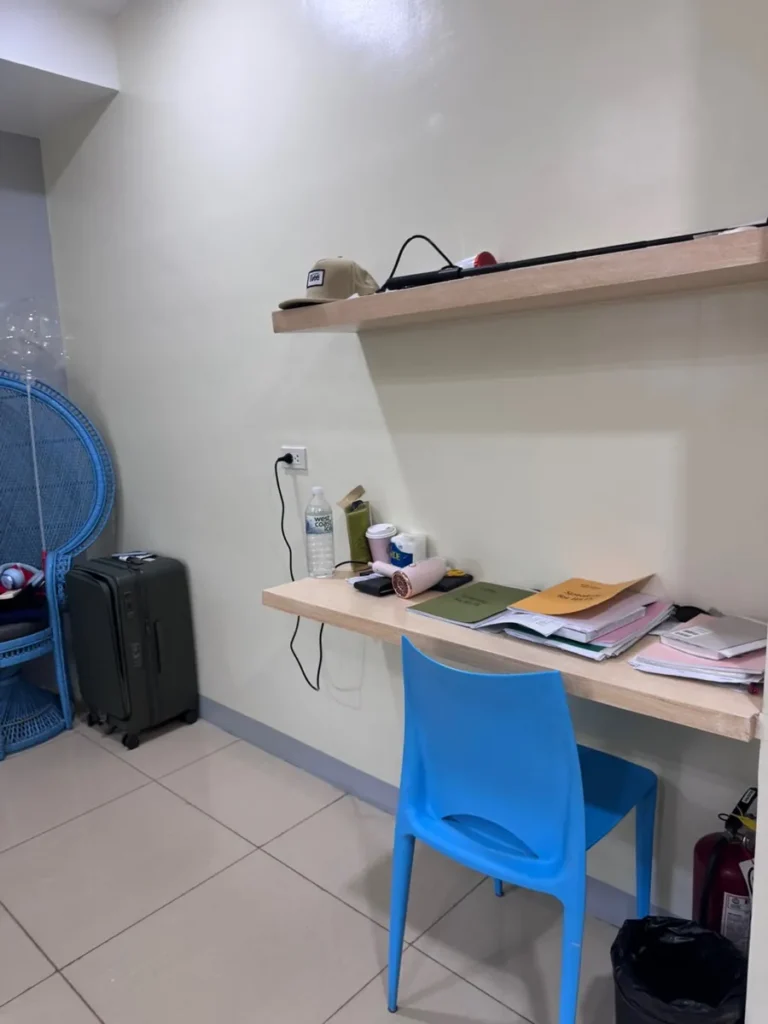 필리핀 세부 셀라(CELLA) 유니캠퍼스 어학원 기숙사 1인실로, 원목 책상과 라탄 의자, 캐리어가 놓인 학습·수납 공간입니다.