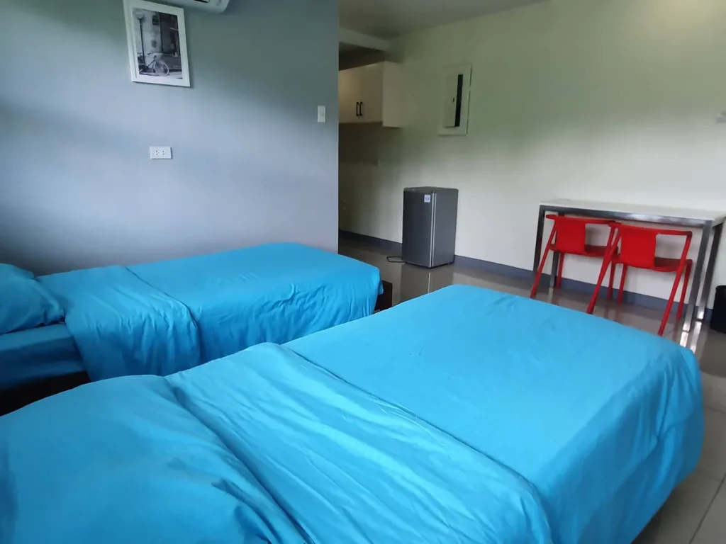 필리핀 세부 셀라(CELLA) 유니캠퍼스 어학원 기숙사 2인실로, 침대 뒤편으로 미니 냉장고와 수납장, 빨간 의자 두 개가 놓인 스틸 책상이 보입니다.