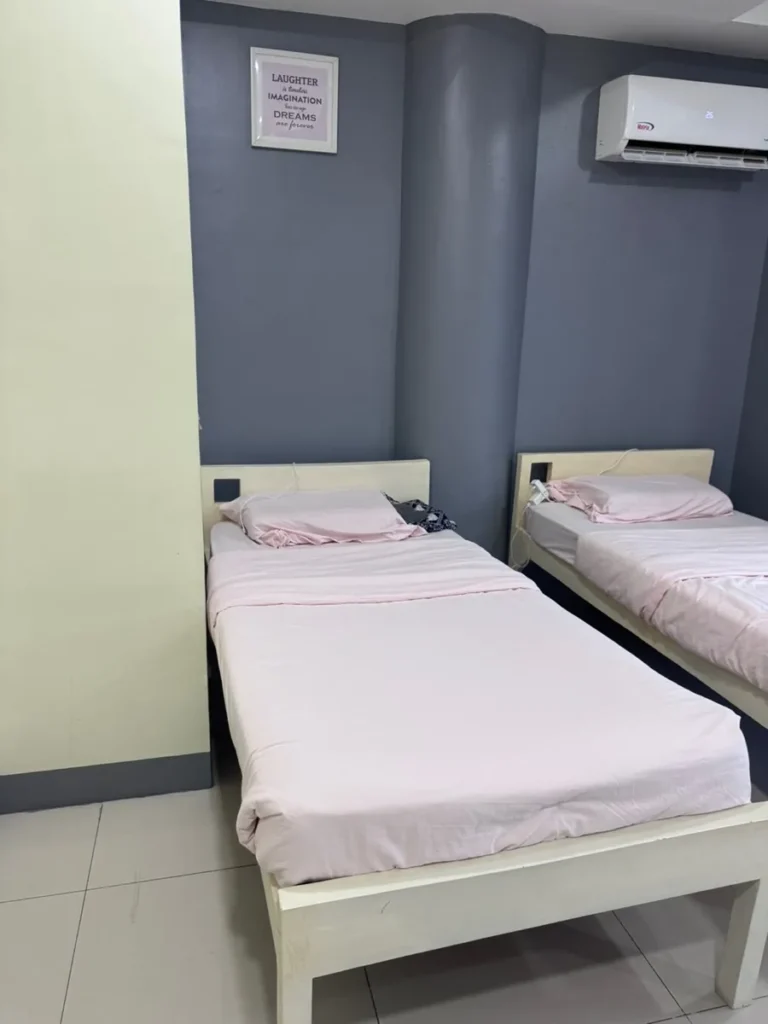 필리핀 세부 셀라(CELLA) 유니캠퍼스 어학원 기숙사 2인실로, 회색 포인트 벽에 액자와 벽걸이 에어컨을 갖춘 분홍 침구의 트윈 베드입니다.