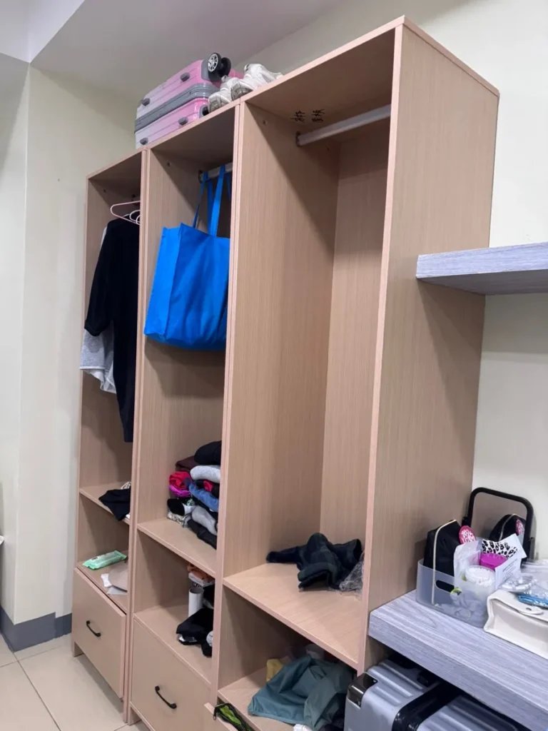 필리핀 세부 셀라(CELLA) 유니캠퍼스 어학원 기숙사 3인실로, 옷걸이에 걸린 옷과 파란 에코백·캐리어가 놓인 개방형 원목 옷장 수납공간입니다.