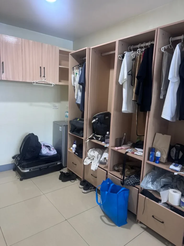 필리핀 세부 셀라(CELLA) 유니캠퍼스 어학원 기숙사 3인실로, 흰 티셔츠와 셔츠들이 걸린 옷장과 상부 벽 캐비닛·캐리어가 보이는 수납공간입니다.