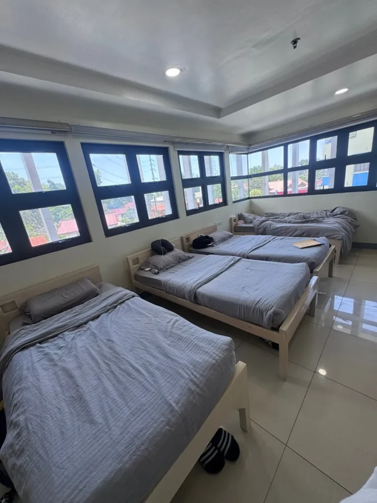 필리핀 세부 셀라(CELLA) 유니캠퍼스 어학원 기숙사 4인실로, 큰 창문으로 햇살이 들어오는 밝은 침실에 원목 침대 세 개가 놓여 있습니다.
