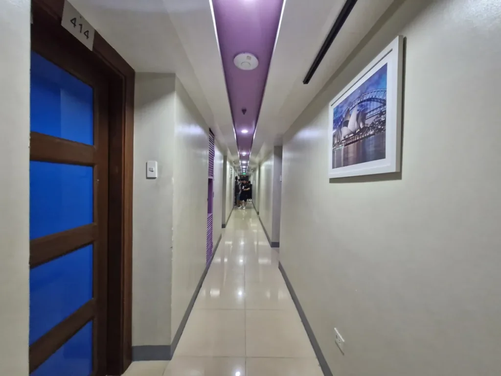 필리핀 세부 셀라(CELLA) 유니캠퍼스 어학원 기숙사 복도로, 보라색 포인트 조명 천장 아래 액자가 걸려 있고 파란 유리문 객실들이 늘어선 복도입니다.