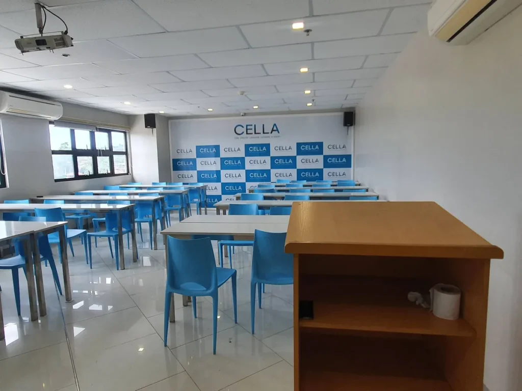 필리핀 세부 셀라(CELLA) 유니캠퍼스 어학원 강의실 컨퍼런스룸으로, 나무 연단 뒤로 파란 의자와 흰 테이블이 정렬된 전경입니다.