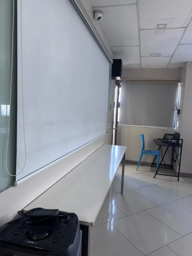 필리핀 세부 셀라(CELLA) 유니캠퍼스 어학원 강의실 컨퍼런스룸으로, 내려진 스크린 옆으로 파란 의자와 장비가 놓인 창가 자리 모습입니다.
