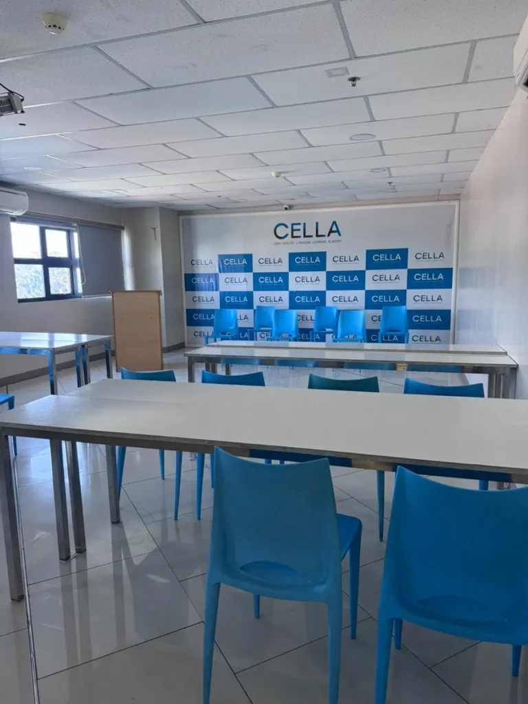 필리핀 세부 셀라(CELLA) 유니캠퍼스 어학원 강의실 컨퍼런스룸으로, CELLA 배경막 앞에 파란 의자와 흰 테이블이 줄지어 놓인 모습입니다.