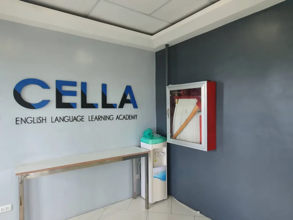 필리핀 세부 셀라(CELLA) 유니캠퍼스 어학원 강의실 복도로, CELLA 학원 로고 벽 옆 정수기와 빨간 소화전함이 놓인 복도 초입입니다.