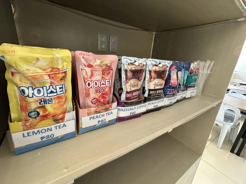 필리핀 세부 셀라(CELLA) 유니캠퍼스 어학원 부대시설 매점으로, 파우치 음료 진열대 옆으로 테이블과 의자가 놓인 매점 내부 전경입니다.