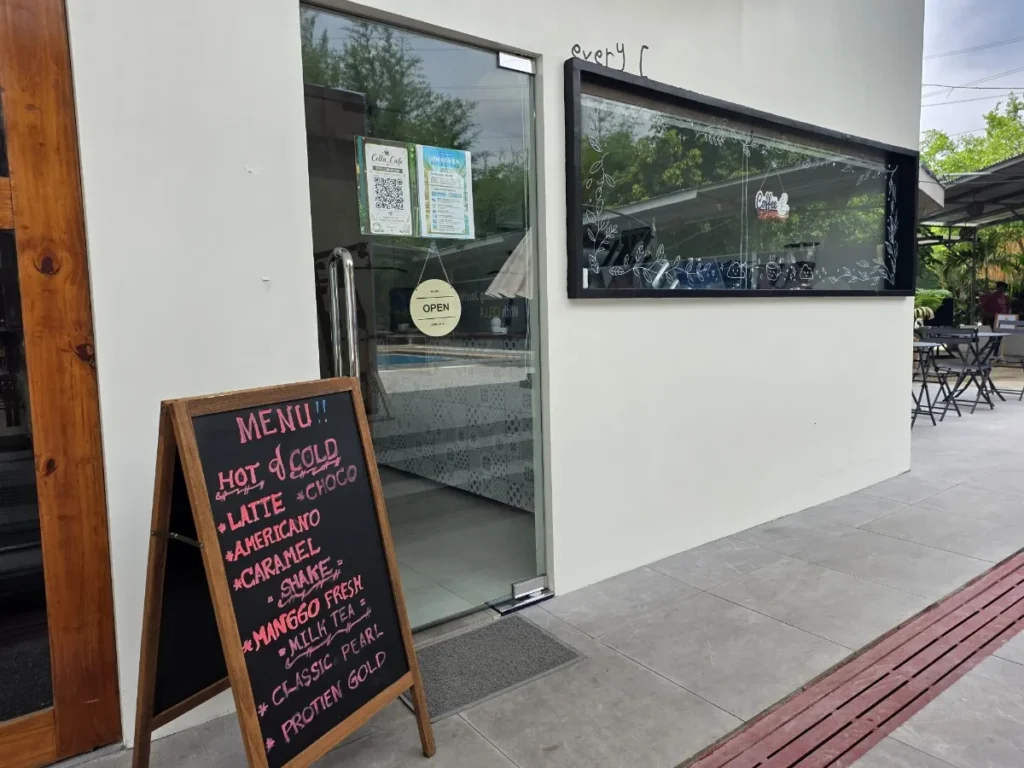 필리핀 세부 셀라(CELLA) 유니캠퍼스 어학원 부대시설 카페로, OPEN 사인과 손글씨 메뉴판이 놓인 유리문 입구에 커피·밀크티 메뉴가 적혀 있습니다.