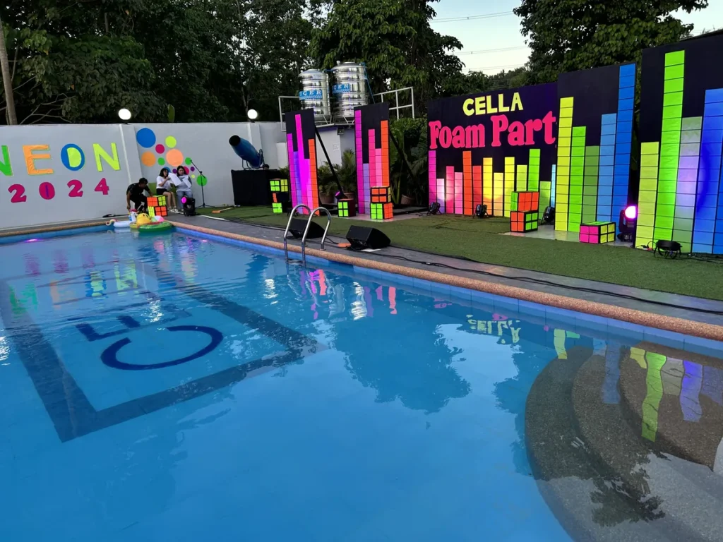 필리핀 세부 셀라(CELLA) 유니캠퍼스 어학원 운동시설 수영장으로, 형광 조명 큐브 장식과 파티 배너 아래 수영장에 로고가 비친 야간입니다.