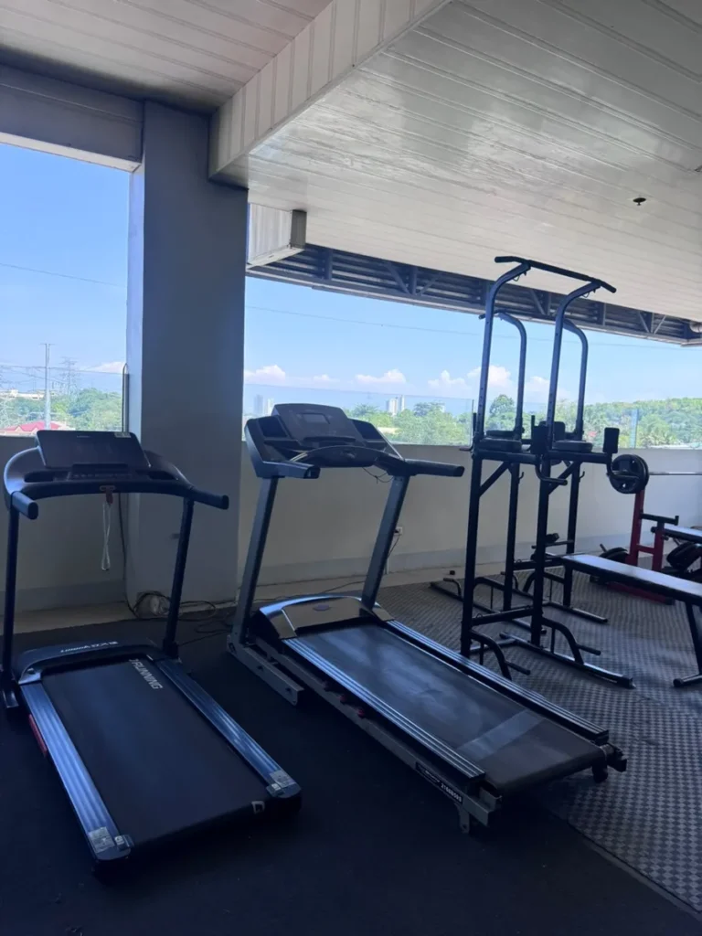 필리핀 세부 셀라(CELLA) 유니캠퍼스 어학원 운동시설 헬스장으로, 도시 스카이라인이 보이는 창가에 트레드밀 두 대와 풀업 파워타워가 놓여 있습니다.