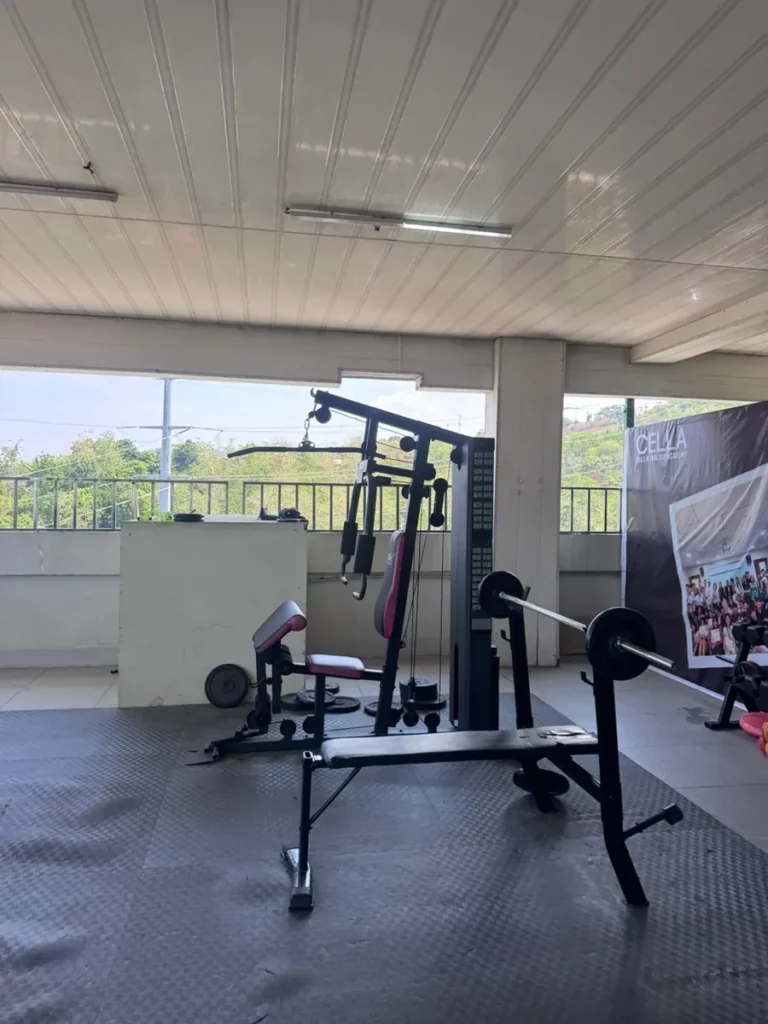 필리핀 세부 셀라(CELLA) 유니캠퍼스 어학원 운동시설 헬스장으로, 초록 나무 전망과 브랜드 배너 옆에 멀티짐과 벤치프레스가 놓인 루프탑입니다.