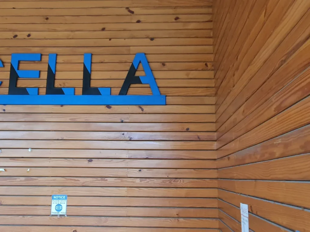 필리핀 세부 셀라(CELLA) 유니캠퍼스 어학원 학원전경 입구로비로, 원목 벽에 부착된 CELLA 로고와 마스크 착용 안내문을 가까이서 담은 모습입니다.