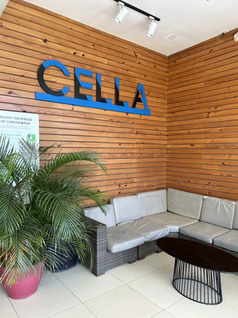 필리핀 세부 셀라(CELLA) 유니캠퍼스 어학원 학원전경 입구로비로, 원목 벽 CELLA 로고 아래 라탄 소파와 야자수 화분이 놓인 로비 코너입니다.