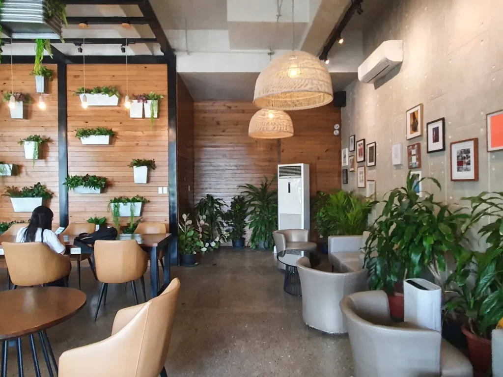 필리핀 세부 셀라(CELLA) 유니캠퍼스 어학원 주변시설 카페로, 원목 화분벽 아래 가죽 의자와 원형 테이블에서 노트북 하는 학생 좌석입니다.