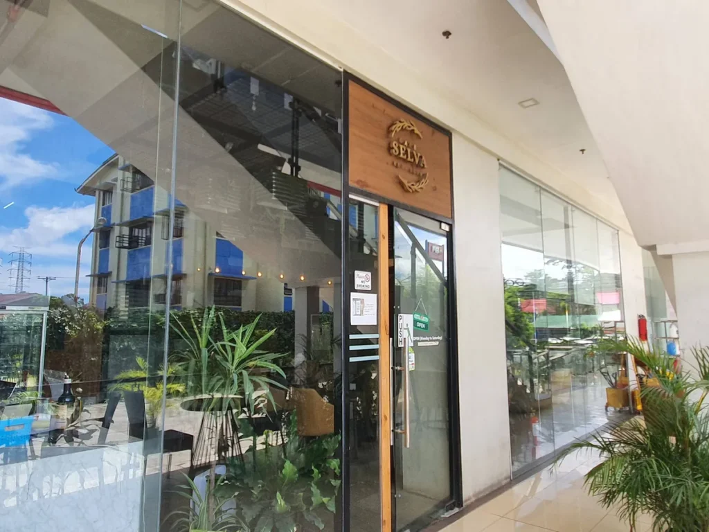 필리핀 세부 셀라(CELLA) 유니캠퍼스 어학원 주변시설 카페로, 원목 간판과 화분이 놓인 통유리 외관, OPEN 표지가 붙은 카페 셀바 입구입니다.