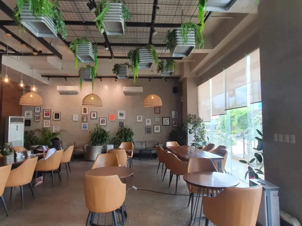 필리핀 세부 셀라(CELLA) 유니캠퍼스 어학원 주변시설 카페로, 라탄 펜던트 조명과 액자 갤러리 벽 아래 원목 테이블 좌석이 늘어선 홀입니다.