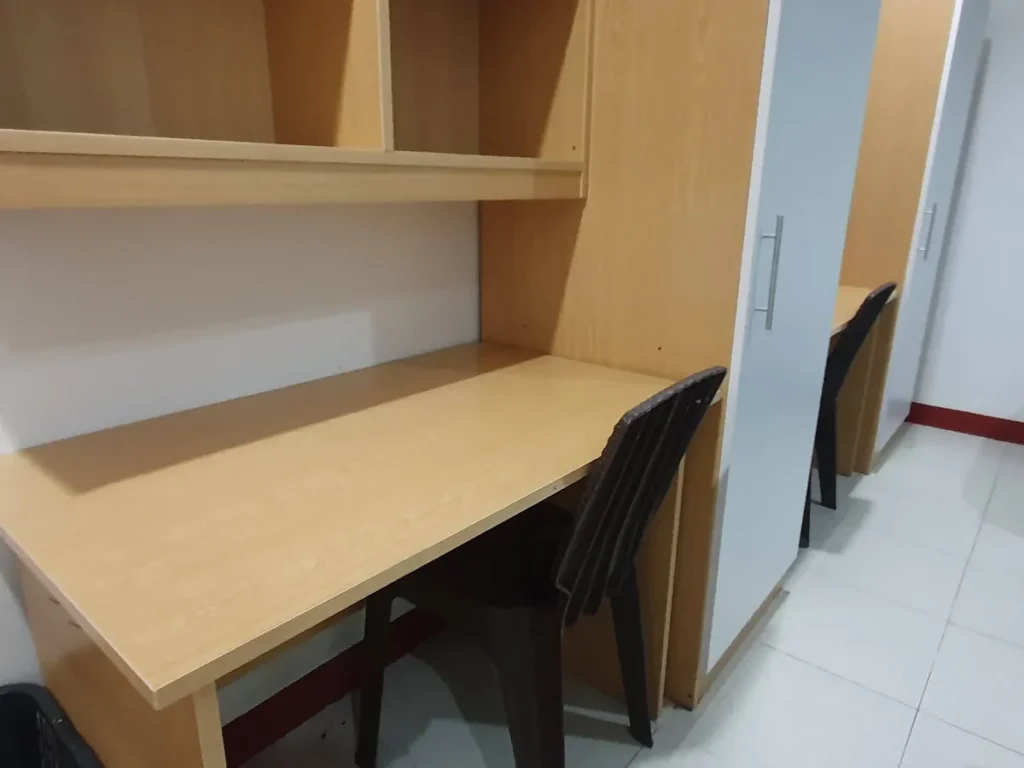 필리핀 세부 CG 바닐라드 어학원 기숙사 2인실로, 원목 책상 위 빈 선반과 옷장 손잡이가 가까이 보이고 검정 철제 의자가 책상 아래 놓인 디테일입니다.